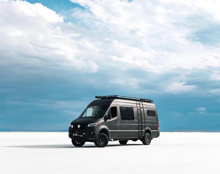 Black van with roof rack on a white surface under a blue sky with clouds